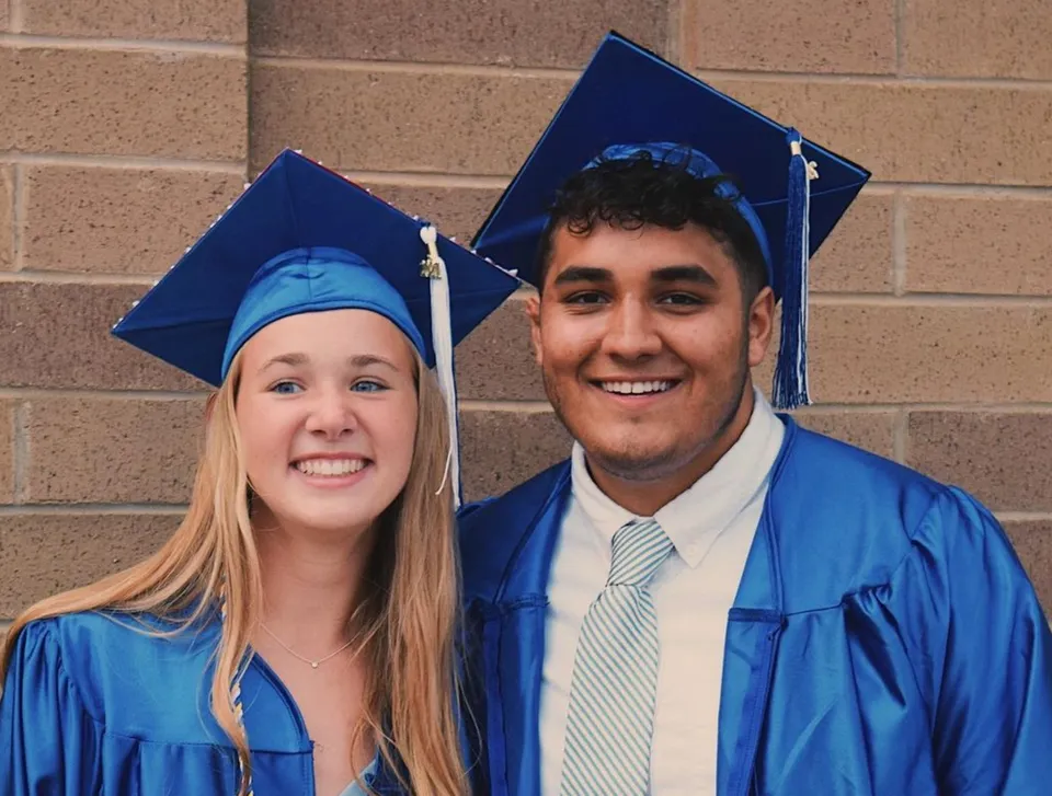 Due neodiplomati sorridenti della Shore Regional High School in toga blu davanti a un muro di mattoni.