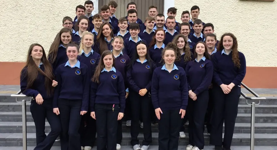 Un gruppo di studenti e studentesse in divise uniformi blu scuro sta davanti a una parete bianca nell'area della Carrigallen Vocational School.