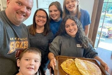 Una estudiante sonriente muestra una bandeja con dos tortillas junto a su familia en casa