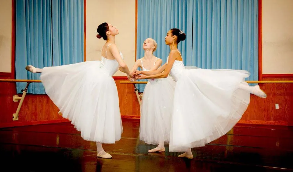 Tre ballerine in tutù bianchi stanno sul pavimento in legno della Northern Academy davanti a tende blu.
