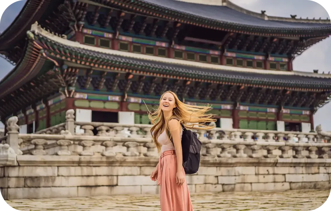 Junge Frau mit Rucksack lacht mit wehenden Haaren vor einem traditionellen koreanischen Palastgebäude mit mehrstöckigem Dach