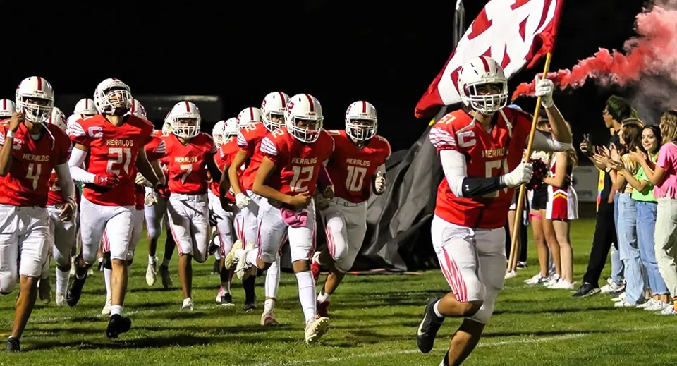 I giocatori di football della Whittier Christian High School corrono insieme sul campo da gioco, mentre sullo sfondo si vedono cheerleader e spettatori.