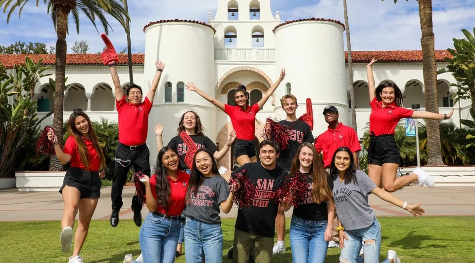 Un gruppo di giovani studentesse in maglie rosse posa davanti a un grande edificio bianco con torre campanaria nel campus della San Diego State University.