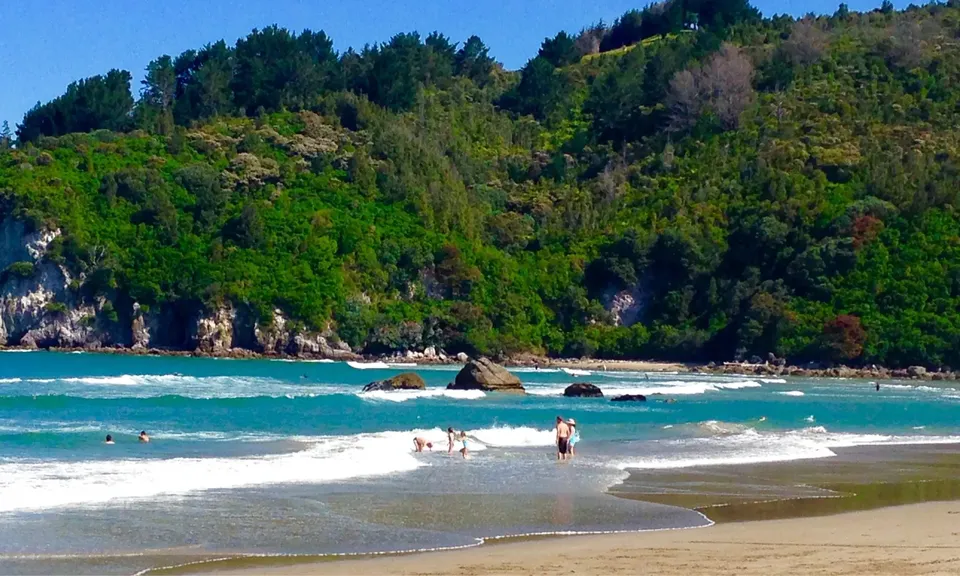 Persone in spiaggia con vista sul mare turchese e sulle colline boscose della zona costiera vicino alla Whangamata Area School.