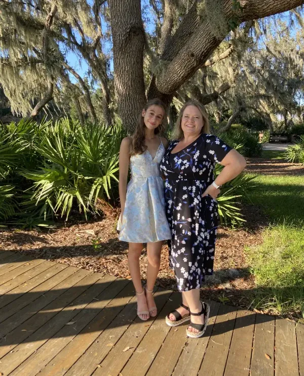 Estudiante con vestido de fiesta junto a su madre anfitriona en un parque bajo un gran árbol