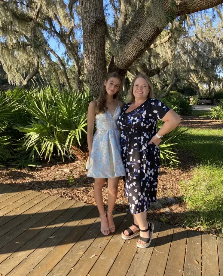Estudiante con vestido de fiesta junto a su madre anfitriona en un parque bajo un gran árbol