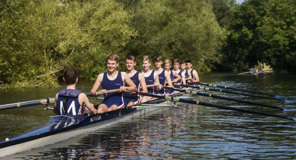 Un gruppo di studenti della Monkton Combe School siede su una barca da canottaggio lungo un fiume tranquillo, con alberi verdi sullo sfondo.