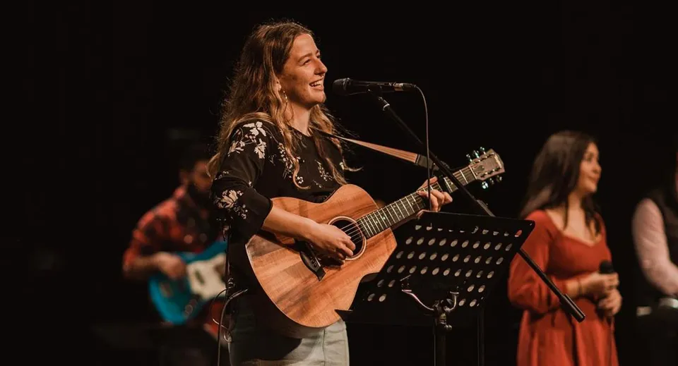 Una studentessa della Spectrum Community Secondary School suona la chitarra acustica su un palco insieme ad altri musicisti.