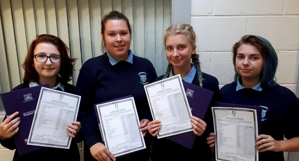 Quattro studentesse in divisa scolastica tengono in mano degli attestati davanti a una parete con tende verticali nel campus del Templemichael College.