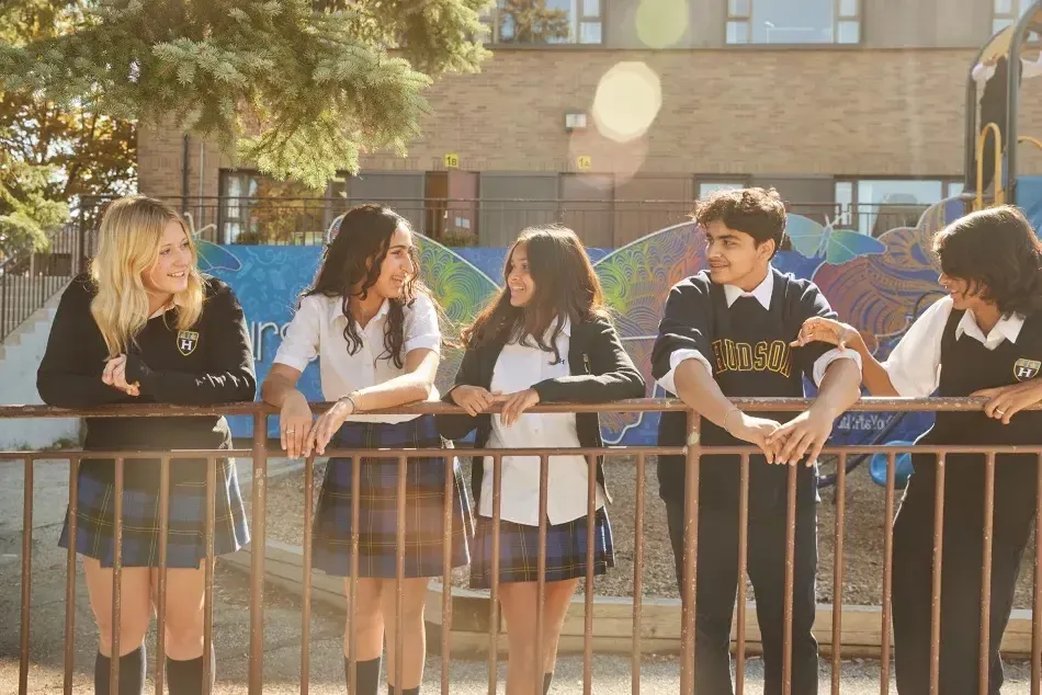 Un grupo de estudiantes está sentado en un banco en la zona exterior del Hudson College con árboles y edificios al fondo.