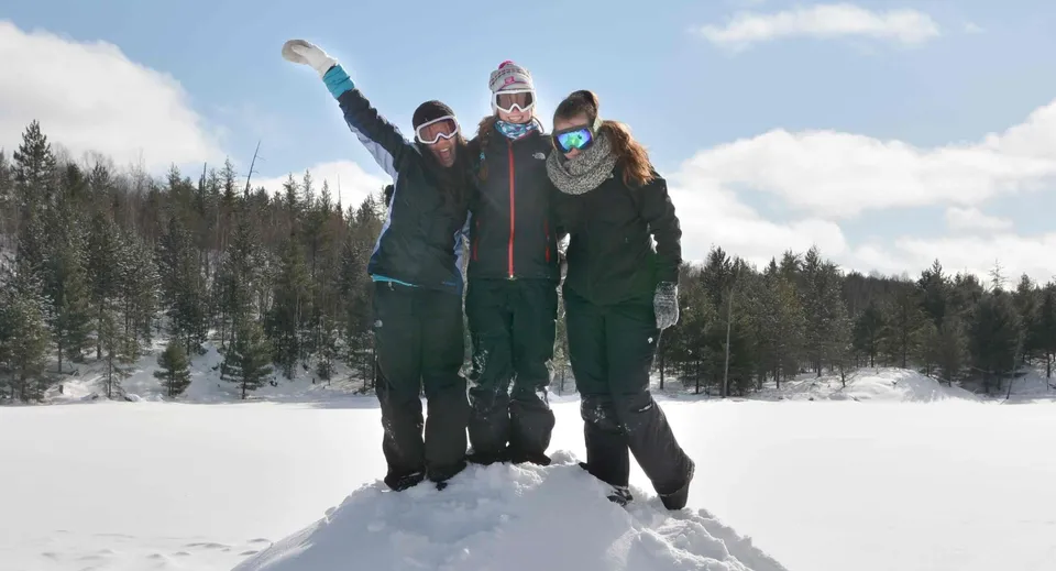 Tre persone in caldi abiti invernali stanno in piedi con le braccia alzate in un bosco innevato nel campus dell'Appleby College.