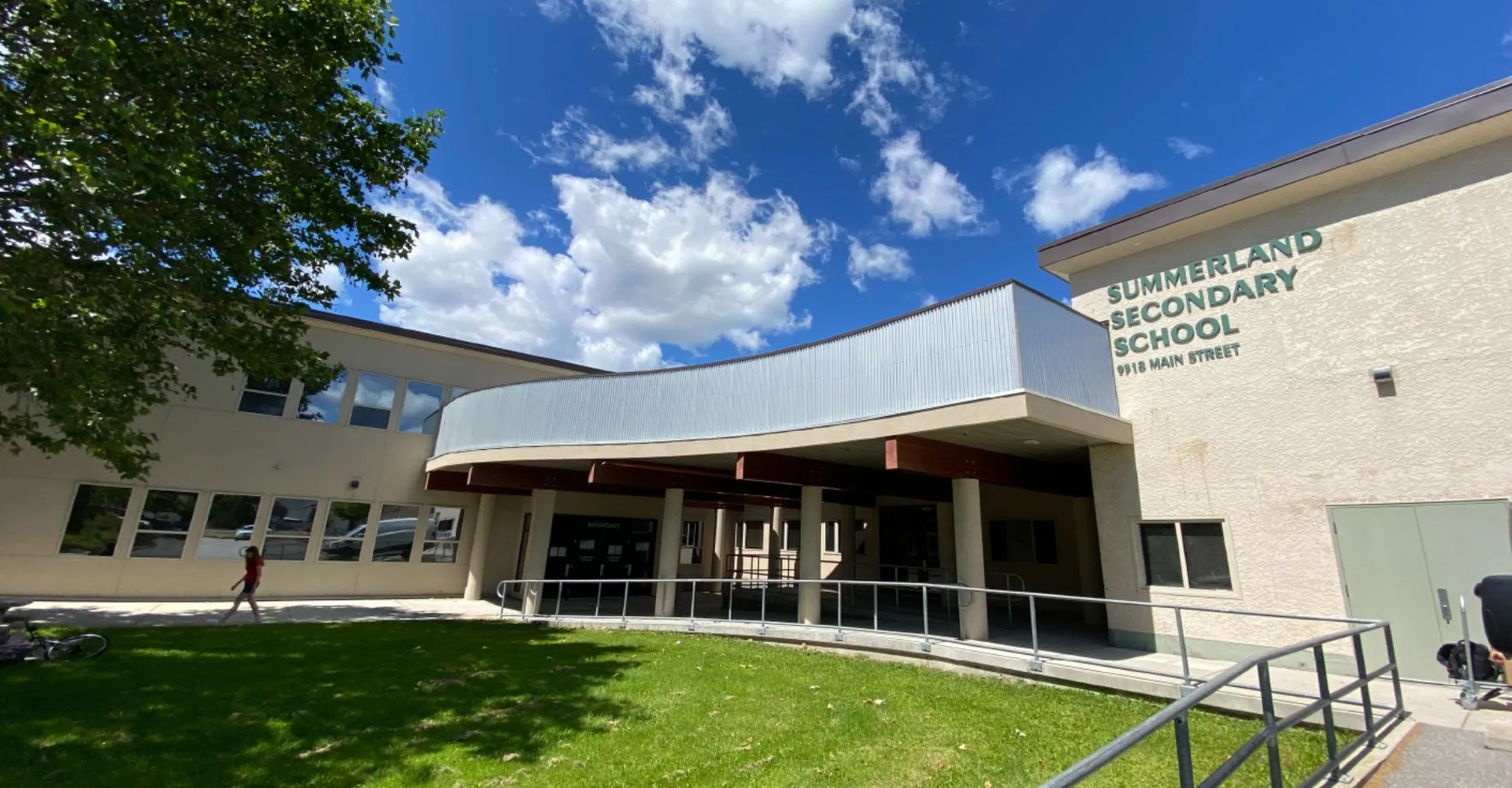 L'edificio moderno a due piani della Summerland Secondary School con ingresso curvo e prato in primo piano sotto un cielo blu.