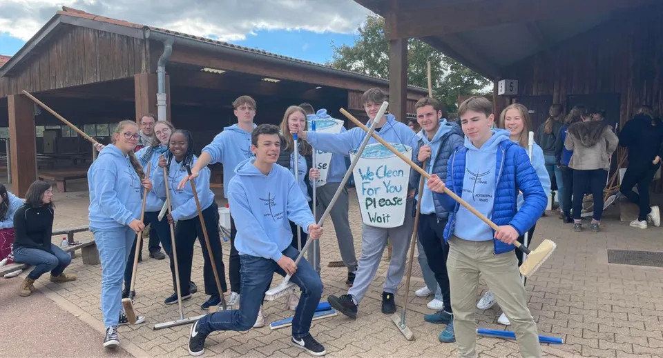 Un gruppo di studenti della Notre Dame Saint Joseph sta in piedi con camicie blu e cartelli davanti a un edificio in legno.
