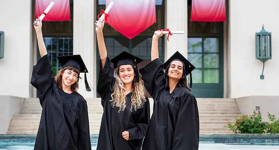Tre studentesse della John Oliver Secondary School sono insieme in abito da cerimonia e tengono con orgoglio i loro diplomi davanti all'edificio scolastico con piscina.