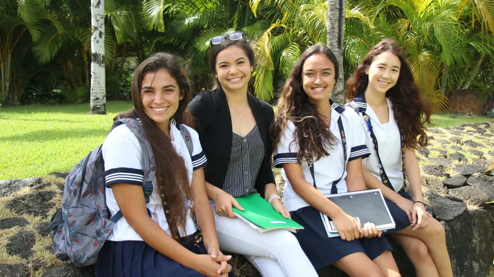 Quattro studentesse della Sacred Hearts Academy sorridono e posano insieme in un giardino tropicale con palme.
