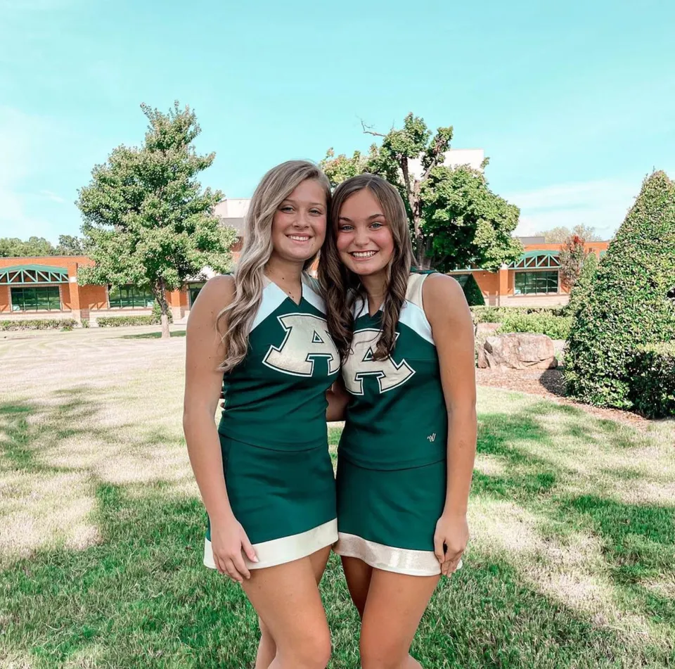 Due studentesse della Alma High School in maglia verde si trovano su un prato con alberi ed edifici sullo sfondo.