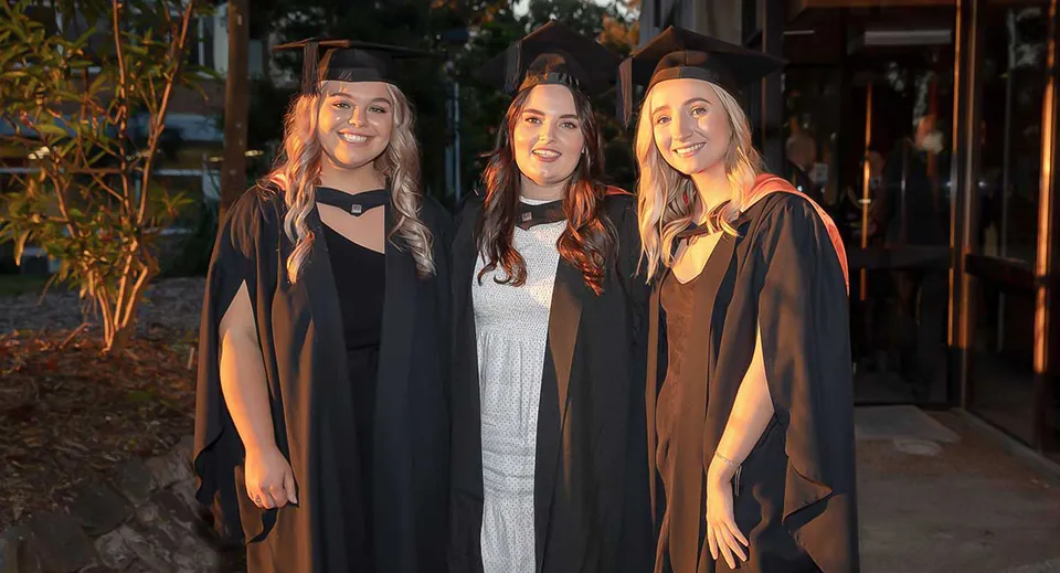 Tre studentesse della Mitchelton State High School in abito da diploma all'aperto davanti ad alberi ed edifici.