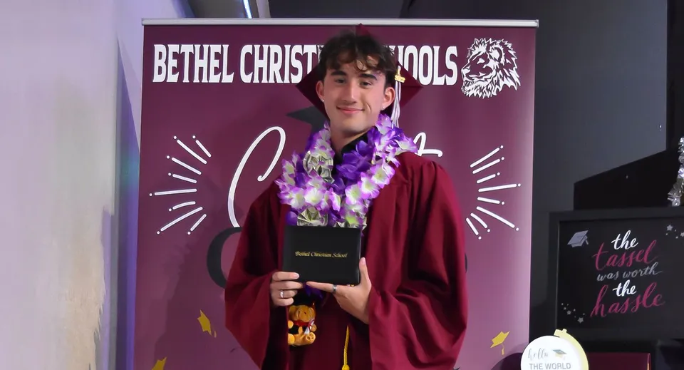 Una persona con una veste rossa e una ghirlanda di fiori sta davanti a uno striscione viola della Bethel Christian Schools con il logo di un leone.
