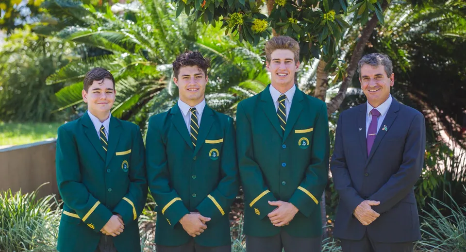 Quattro studenti del St. Brendan's College sono in piedi in abiti verdi e viola in un'area esterna tropicale con palme.