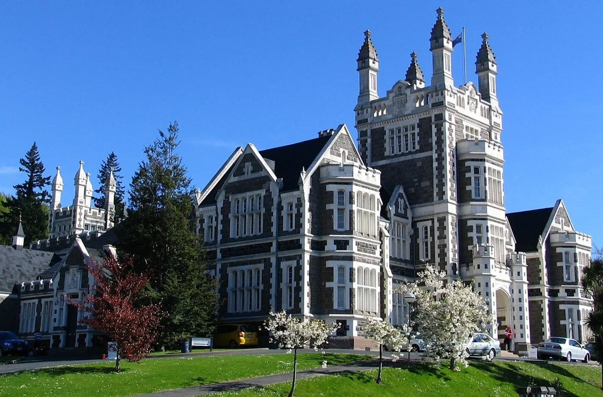 L'edificio principale in stile gotico della Otago Boys' High School è circondato da alberi in fiore sotto un cielo blu.