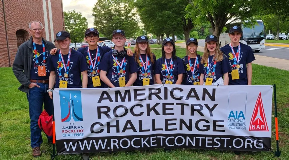Studenti della The Vanguard School in posa davanti a uno striscione della American Rocketry Challenge con alberi ed edifici sullo sfondo.
