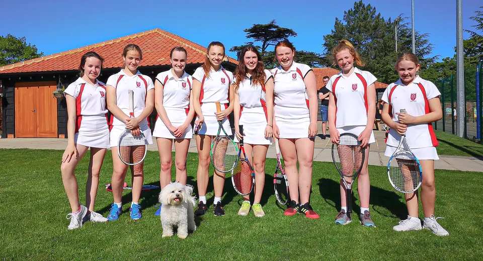 Un gruppo di studenti e studentesse della Royal Hospital School in abbigliamento da tennis sta con un cane su un prato davanti a un edificio con il tetto di tegole rosse.