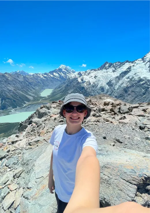 Louisa lächelt auf einem Berggipfel mit schneebedeckten Gipfeln und einem Gletschersee in Neuseeland