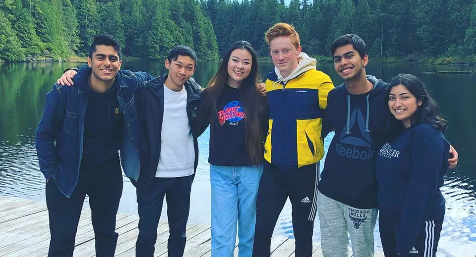 Un gruppo di studenti della Burnaby Mountain School si trova su un pontile in legno davanti a un lago calmo circondato da alberi verdi.