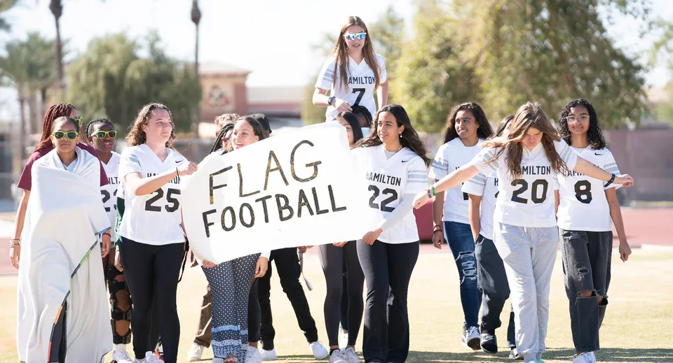 Un gruppo di studentesse della Hamilton High School si trova con un cartello di flag football davanti al campus scolastico.
