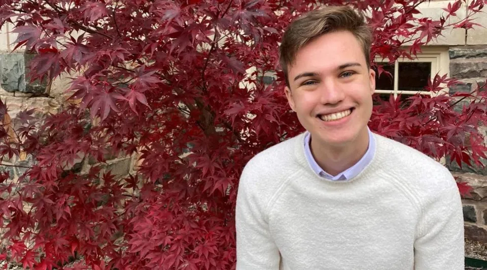 Una persona con un maglione bianco sta davanti a foglie autunnali nel campus della FJ Brennan Catholic High School.