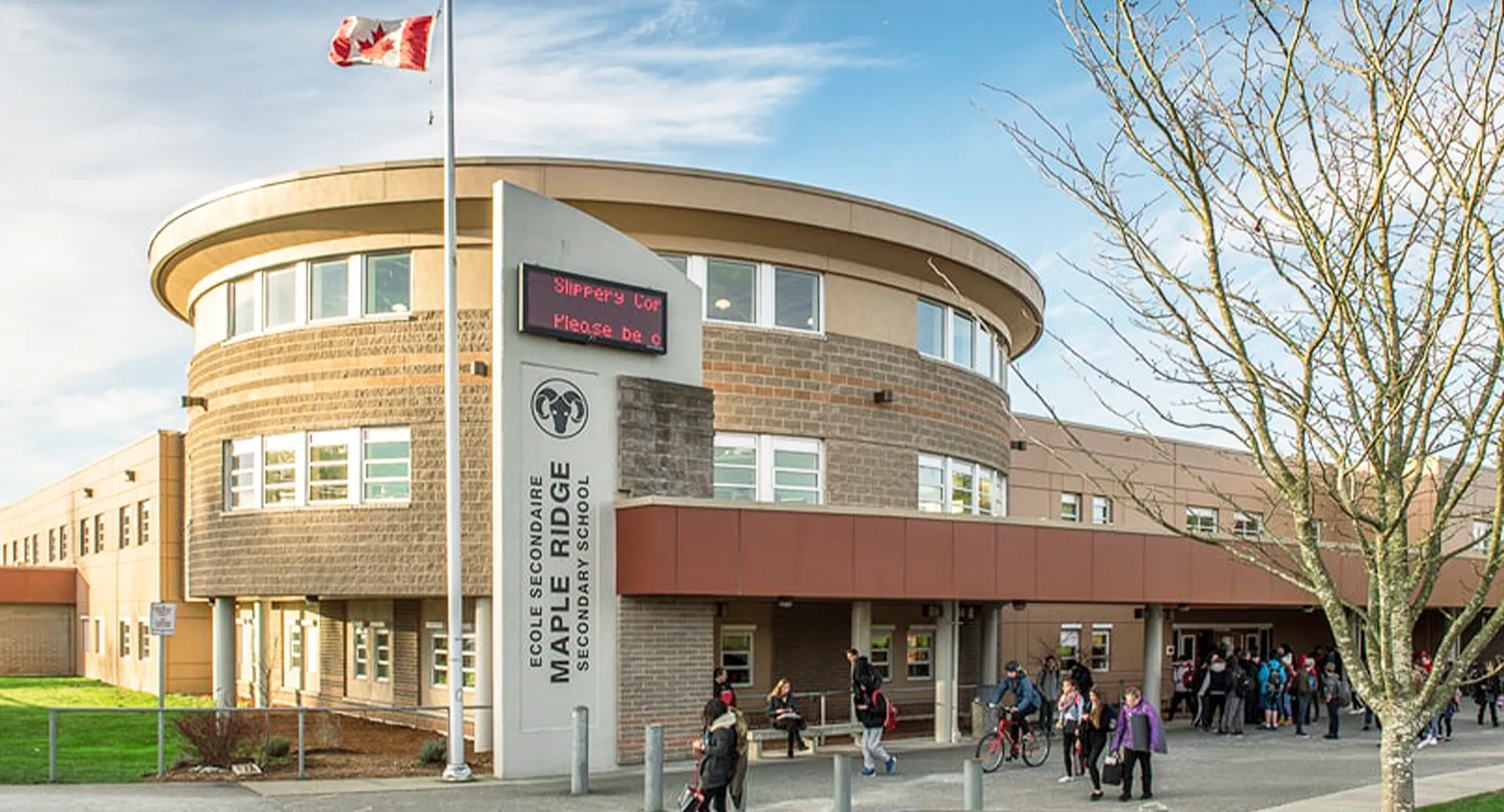 L'edificio moderno e circolare della Maple Ridge Secondary School con una bandiera canadese sul tetto e persone sul marciapiede.