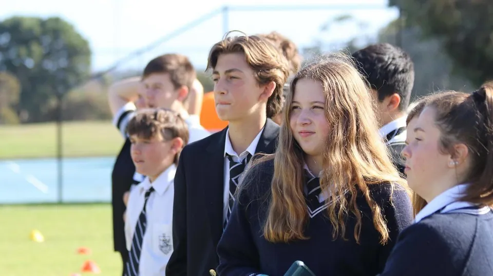 Un gruppo di studenti della Adelaide High School si trova su un campo erboso accanto agli edifici scolastici e agli alberi.