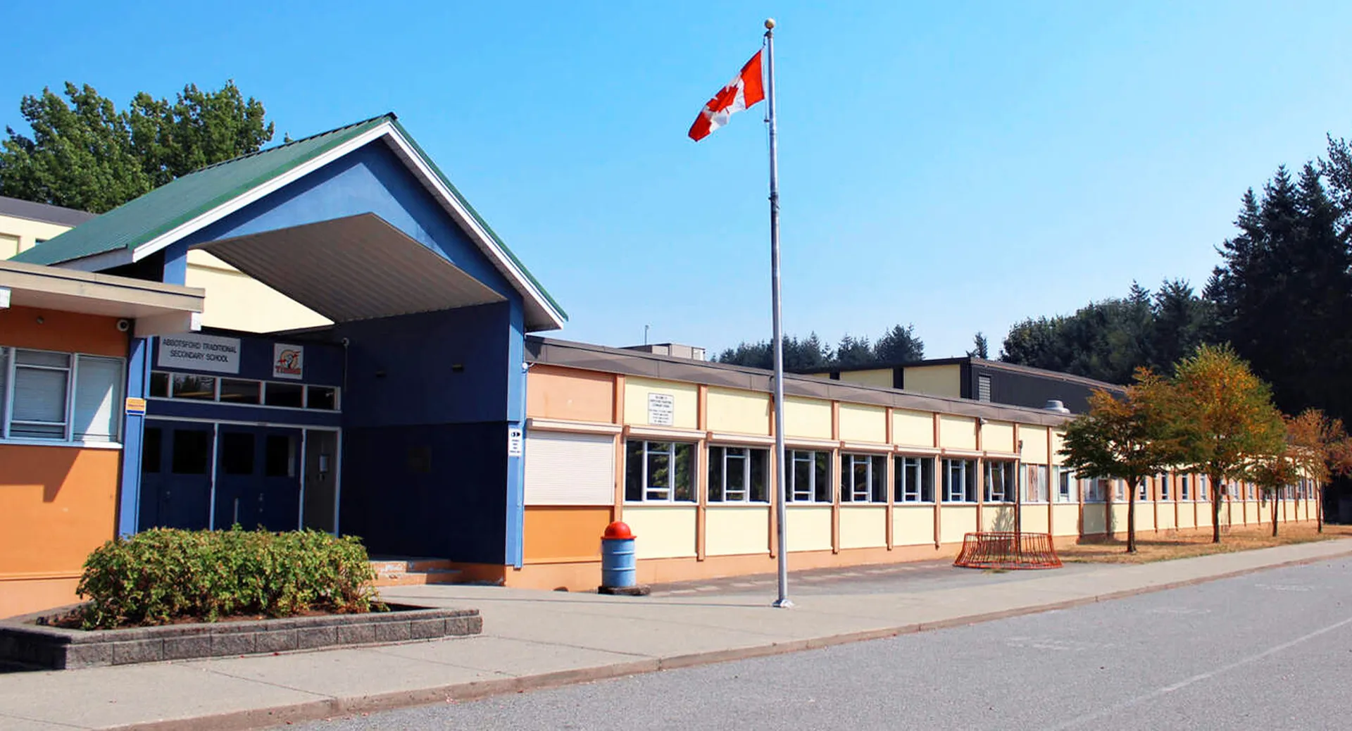 L'edificio moderno a più piani della Abbotsford Traditional Secondary School con una bandiera canadese sul tetto e alberi in primo piano.