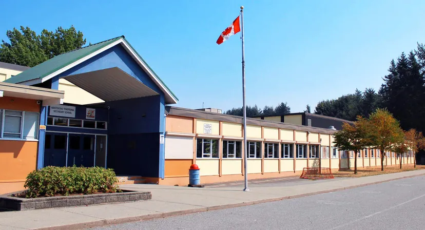 L'edificio moderno a più piani della Abbotsford Traditional Secondary School con una bandiera canadese sul tetto e alberi in primo piano.
