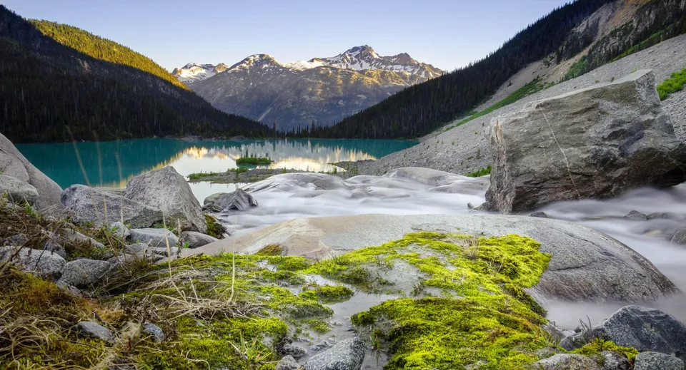 Un lago di montagna turchese in Canada con cime innevate e un ruscello che scorre tra rocce ricoperte di muschio.
