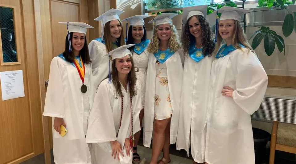 Un gruppo di diplomande della East Catholic High School in toga bianca con medaglie posa insieme nell'edificio scolastico.