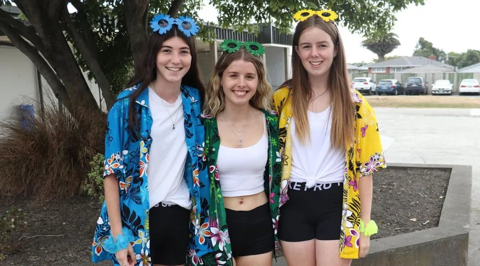 Tre studentesse della Kaiapoi High School posano ridendo all'aperto davanti ad alberi ed edifici.