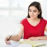 Chica joven con camiseta roja escribiendo con bolígrafo en un cuaderno sobre el escritorio