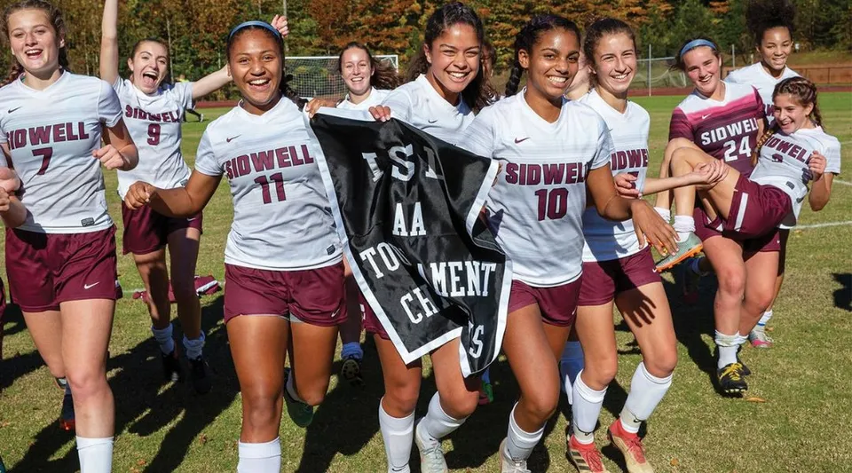 Un gruppo di giovani calciatrici della Sidwell Friends School si raccoglie sorridente su un campo erboso con alberi sullo sfondo.