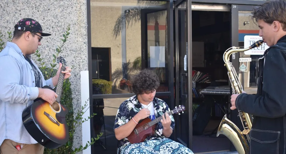 Tre studenti della John F. Kennedy High School suonano la chitarra, l'ukulele e il sassofono davanti a un edificio.