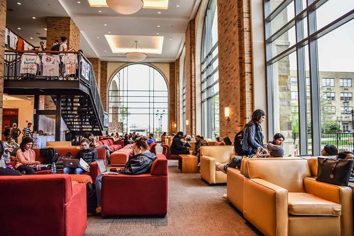 Salón del campus con sillones rojos y naranjas, estudiantes sentados y grandes ventanales