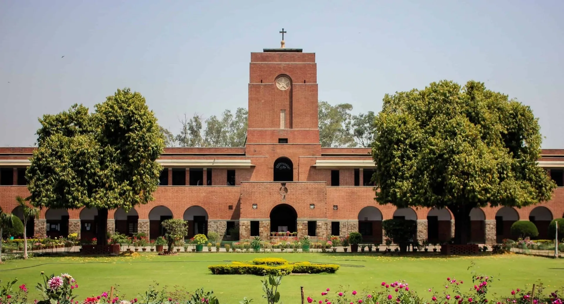 L'edificio in mattoni con un'alta torre e una croce nel campus del Saint Stephen’s College è circondato da alberi verdi e aiuole ben curate.