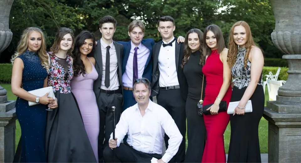Un gruppo di persone ben vestite si trova insieme davanti a uno sfondo verde nel terreno della Monkton Combe School.