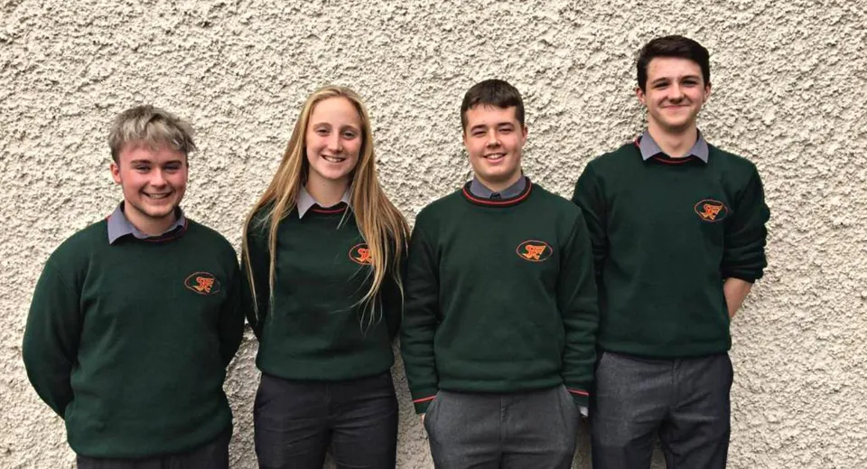 Quattro studenti e studentesse in maglione verde sono in piedi davanti a una parete bianca nel campus del St. Fergal’s College.