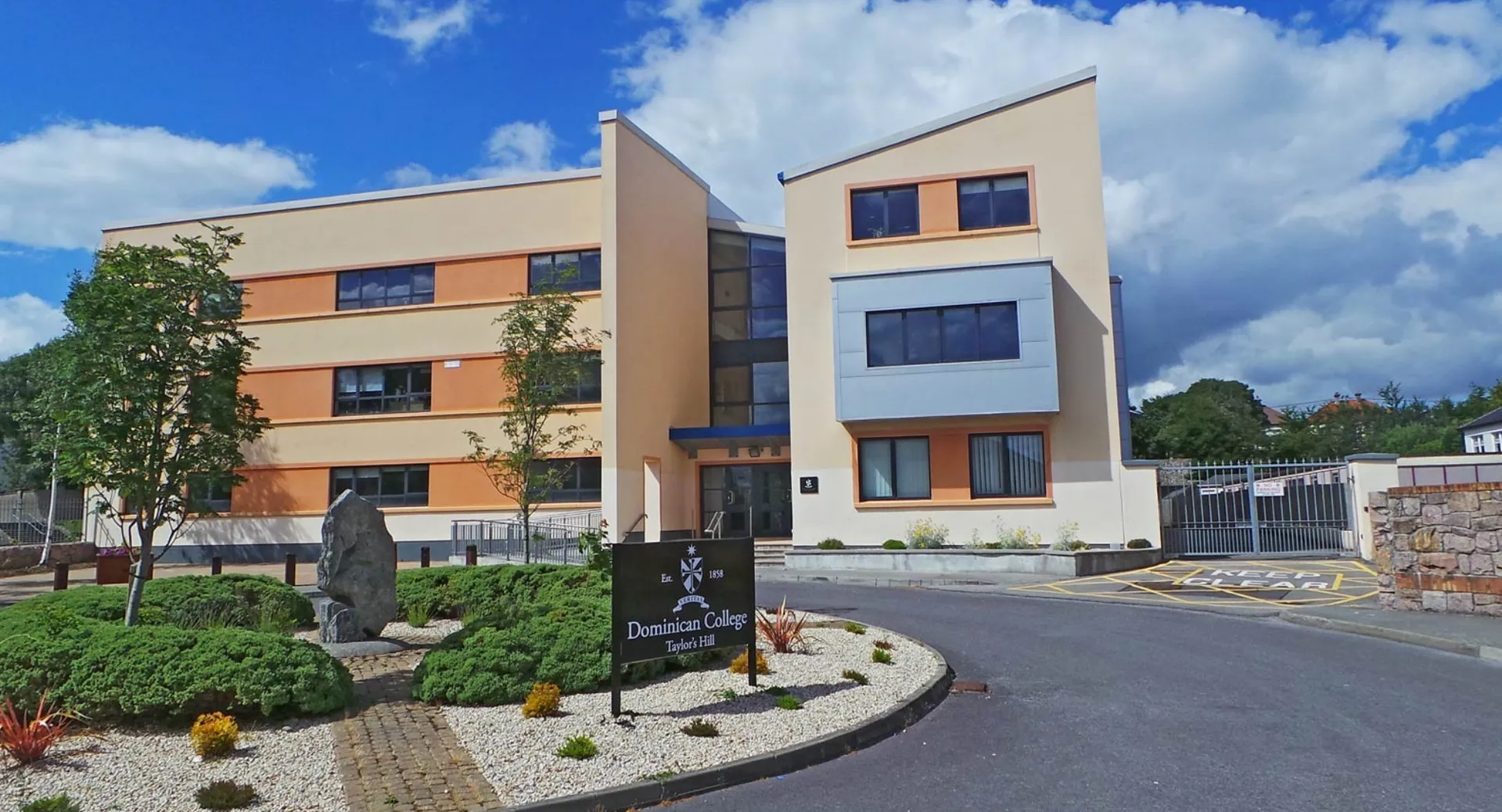 L'edificio moderno del Dominican College Galway con un giardino curato e una scultura davanti a un cielo blu.