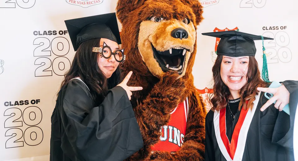 Due studentesse della Britannia Secondary School in toga da diploma posano con la mascotte della scuola davanti allo sfondo Class of 2020.