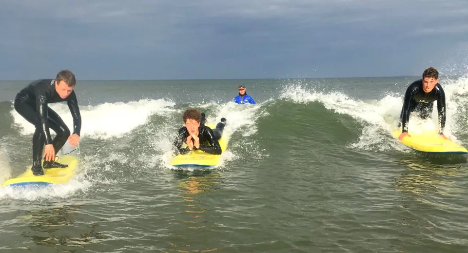Studenti della Royal School Cavan fanno surf in muta sulle onde dell'oceano sotto un cielo nuvoloso.