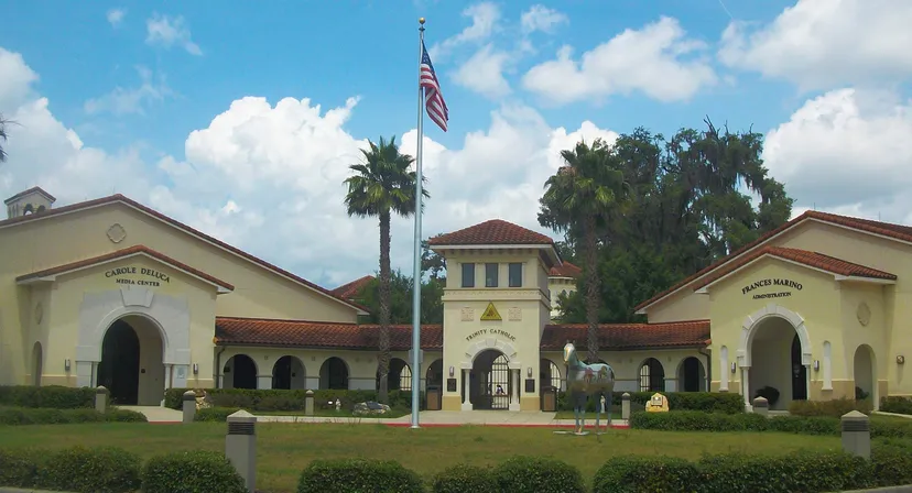 L'edificio principale della Trinity Catholic High School mostra uno stile spagnolo con torre e bandiera americana davanti a palme e cielo blu.