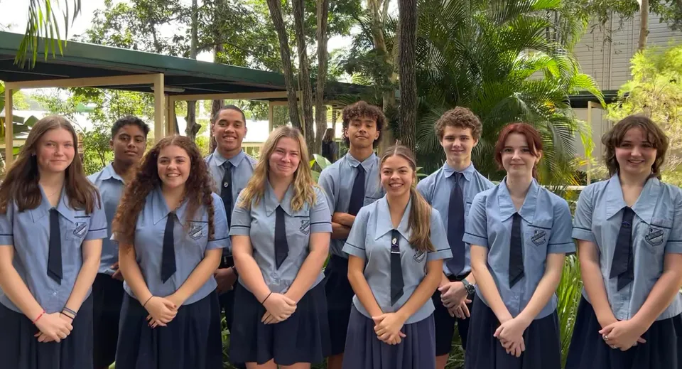 Un gruppo di studentesse e studenti della Holland Park State High School posa insieme in un'area verde all'aperto con alberi ed edifici scolastici sullo sfondo.