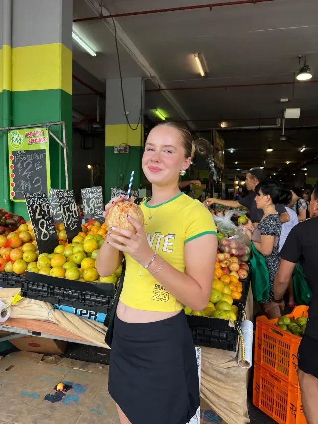 Studentessa con una noce di cocco in mano sorride tra le bancarelle di frutta di un mercato australiano